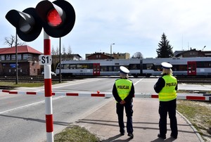 Policjantki w białych czapkach i żółtych odblaskowych kamizelkach stoją przy szlabanie kolejowym. W tle przejeżdża pociąg.