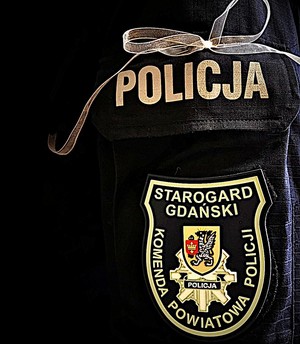 Lewe ramię policjanta, na którym przypięta jest biała wstążka. Poniżej tarcza z logiem Komendy Powiatowej Policji w Starogardzie Gdańskim.