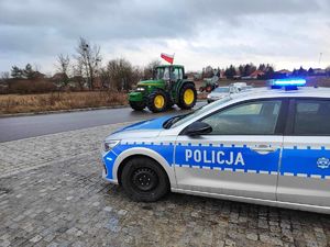 Radiowóz policyjny stoi przy drodze, po której jedzie ciągnik rolniczy.