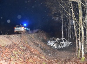 Droga przebiegająca przez las. Po lewej stronie radiowóz z włączonymi błyskami, a po prawej stronie na poboczu rozbite i spalone auto.