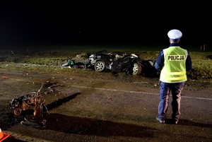 Policjant w żółtej kamizelce odblaskowej i białej czapce stoi przed rozbitym pojazdem. Po lewej stronie leży wyrwany z Chevroleta silnik.