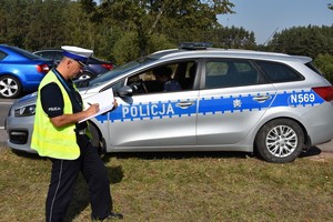 Policjant sprawdza prawidłowość infrastruktury drogowej. W tle oznakowany radiowóz.