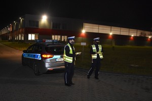 Policjanci trzymają kamizelki odblaskowe oczekując na osoby idące do pracy. W tle Radiowóz oznakowany.