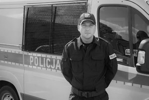 Na pierwszym planie Św. pamięci funkcjonariusz. W tle oznakowany radiowóz policyjny.