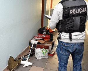 Policjant prowadzi oględziny odzyskanych przedmiotów, które leżą rozłożone przed nim na korytarzu w budynku KPP.