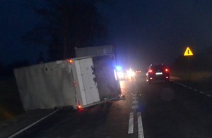 Przewrócona na boku ciężarówka. W oddali radiowóz zabezpieczający miejsce wypadku.