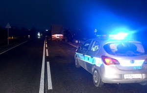 Radiowóz policyjny z włączonymi światłami błyskowymi zabezpiecza miejsce wypadku. W oddali widać rozbite auta.