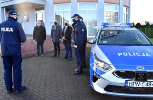 Zdjęcie z uroczystości przekazania nowego radiowozu na tle budynku Komendy Powiatowej Policji w Starogardzie Gdańskim. W górnej lewej części kadru przedstawiciele samorządu podczas uroczystości przekazania radiowozu. W prawej części kadru przekazywany radiowóz.