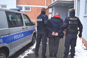 Policjanci prowadzą jednego z podejrzanych. W lewej części zdjęcia oznakowany radiowóz.