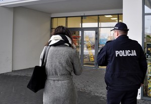 Policjant z pracownicą Sanepidu przed drzwiami kontrolowanej placówki handlowej zmierzają w jej kierunku.