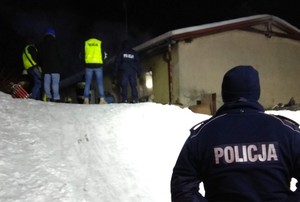 Policjant zabezpiecza miejsce zdarzenia. Funkcjonariusz zwrócony plecami do fotografującego. Na mundurze widoczny biały napis Policja. W tle dogaszany budynek oraz inne pracujące na miejscu służby.