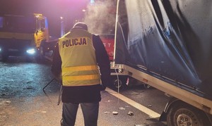 Technik kryminalistyki przeprowadza czynności procesowe na miejscu zdarzenia. W tle pojazdy służb ratowniczych z włączonymi światłami błyskowymi.