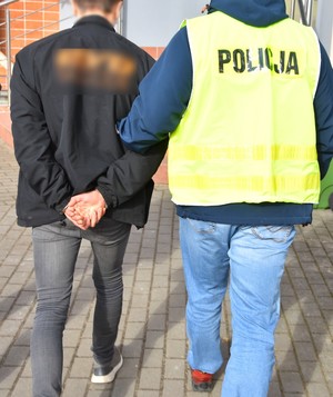 Policjant prowadzi zatrzymanego mężczyznę, który ma założone kajdanki.