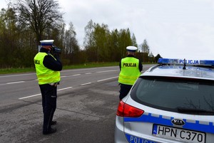 Policjanci stoją przy zaparkowanym przy drodze radiowozie. Jeden z funkcjonariuszy mierzy prędkość nadjeżdżającego auta.