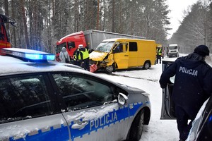 W dolnej części zdjęcia stojący bokiem do widza oznakowany radiowóz policyjny z włączonym kogutem. Nad jego dachem w pewnej odległości stoją rozbite auta, które uczestniczyły w wypadku. Po prawej stronie kadru widać policjanta wsiadającego do radiowozu.