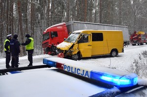W dolnej części zdjęcia stojący bokiem do widza oznakowany radiowóz policyjny z włączonym kogutem. Nad jego dachem w pewnej odległości stoją rozbite auta, które uczestniczyły w wypadku. W lewej części kadru widoczni są policjanci pracujący na miejscu zdarzenia.