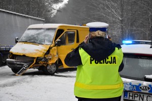 Na pierwszym planie stoi policjantka zwrócona do widza plecami. Policjantka przygląda się uszkodzeniom stojącego przed nią rozbitego żółtego mercedesa.