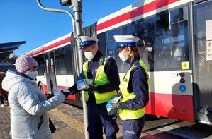 Policjanci na przystanku autobusowym wręczają kobiecie maseczkę.