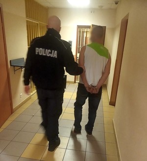 Policjant na korytarzu komendy prowadzi zatrzymanego, który ma założone kajdanki.