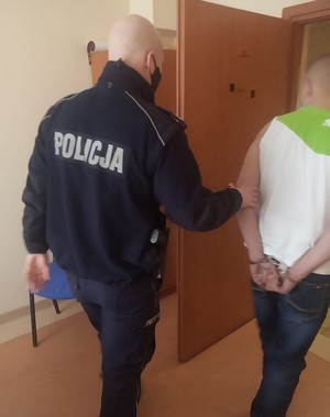 Policjant wyprowadza podejrzanego z biura po przesłuchaniu. Podejrzany ma założone kajdanki na ręce trzymane z tyłu.