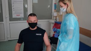 Policjant w gabinecie lekarskim oczekuje na szczepienie. Po jego prawej stronie stoi pielęgniarka.