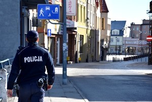 Umundurowany policjant zwrócony do obiektywu plecami podczas patrolu pieszego. W tle znaki drogowe i zabudowania ulicy Chojnickiej.