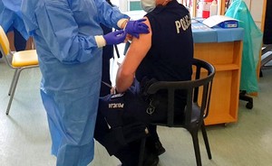 Policjant siedzi na krześle, a po jego lewej stronie stoi pielęgniarka trzymająca igłę.