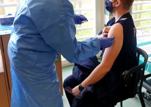 Policjant siedzi na krześle, a po jego lewej stronie stoi pielęgniarka trzymająca igłę.