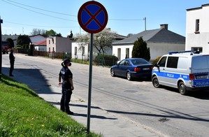 Stojący na chodniku policjanci zabezpieczają strefę zagrożoną przed dostępem osób postronnych. W tle zaparkowany radiowóz policyjny.