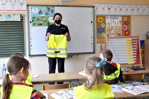 Policjantka stoi przy tablicy i prezentuje dzieciom siedzących w ławce kamizelkę odblaskową.