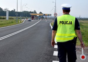 Policjant stoi przy bramkach autostrady A1. W prawej dłoni trzyma policyjnego lizaka.