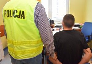 Policjant stoi przy zatrzymanym mężczyźnie, który siedząc na krześle oczekuje na przesłuchanie.
