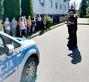 Policjanci rozmawiają w plenerze z uczniami o bezpiecznych zachowaniach w czasie wakacji.