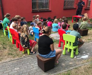 Dzieci siedzą na krzesełkach i słuchają wypowiedzi policjanta.