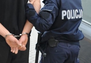 Policjant prowadzi zatrzymanego mężczyznę, który ma założone kajdanki na rękach trzymanych z tyłu.