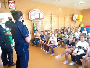 Policjanci podczas prelekcji dla uczniów młodszych klas. Dzieci siedzą na ławeczkach, a na przeciwko stoją policjanci prowadzący zajęcia.
