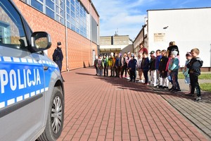 Policjantka stoi naprzeciwko grupy dzieci. po lewej stronie kadru zaparkowany jest radiowóz policyjny.