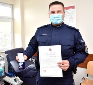 Uhonorowany policjant w mundurze pokazuje medal oraz otrzymany dyplom. W tle fotel do pobierania krwi.