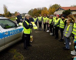 Policjantka rozmawia w plenerze z grupą uczniów przed egzaminem. W tle oznakowany radiowóz policyjny.