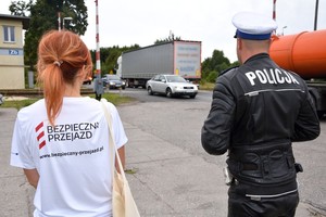 Policjant z pracownicą kolei obserwują pojazdy przejeżdżające przez przejazd kolejowy.