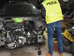 Policjant prowadzi oględziny odzyskanego pojazdu. Auto ma całkowicie rozebrany przód.