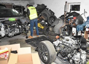Policjant prowadzi oględziny odzyskanego pojazdu. Przód pojazdu całkowicie rozkręcony. W pobliżu auta znajduje się sporo części samochodowych.