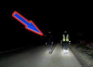 Zdjęcie obrazuje widoczność pieszych na drodze po zmierzchu. Obok siebie stoją dwie osoby, a tylko jedna posiada kamizelkę odblaskową. Druga osoba jest prawie niewidoczna w świetle samochodowych reflektorów.