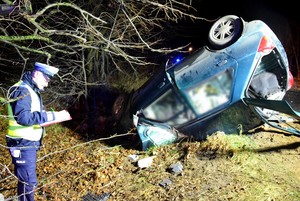 Policjant prowadzi oględziny rozbitego forda. Auto po dachowaniu leży na dachu.