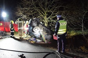 Służby pracują na miejscu wypadku. Na pierwszym planie widoczny policjant w żółtej kamizelce, dalej rozbite auto oraz ratownicy straży pożarnej.