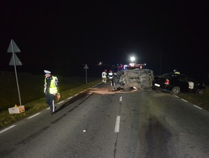 Policjanci z drogówki pracują na miejscu wypadku. W tle rozbite auta osobowe.