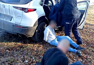 Policjant przeszukuje samochód, przy którym na ziemi siedzą zakuci w kajdanki podejrzani.