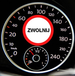 Grafika obrazkowa. Tarcza prędkościomierza bez wskazówki. W środku tarczy znajduje się znak drogowy z kategorii znaków zakazu z czarnym napisem o treści zwolnij.