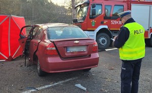 Zdjęcie rozbitego chevroleta. W pobliżu policjant drogówki prowadzi czynności służbowe.