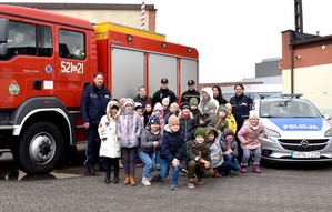 Zdjęcie grupowe na placu apelowym. Policjanci, strażacy oraz dzieci. Po bokach pojazdy służbowe wymienionych służb.